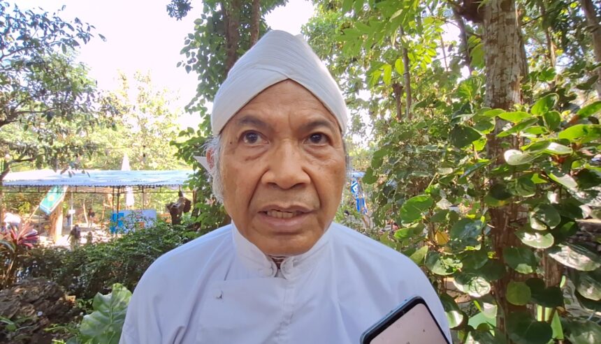 Close-up of an elderly man in a white turban outdoors, with greenery in the background and a smartphone visible in the foreground.