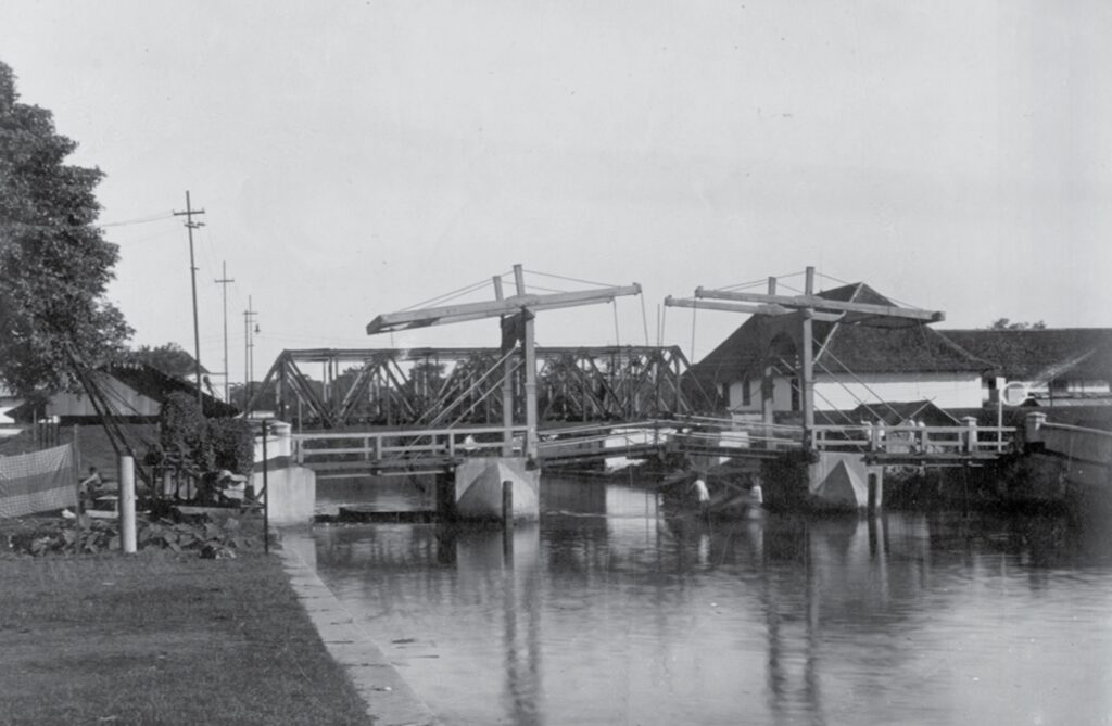 Screenshot_20260610_020204_WPS Office - Mabur.co Historic canal scene with a large metal drawbridge spanning the water and timber supports, flanked by houses and a tree-lined bank.