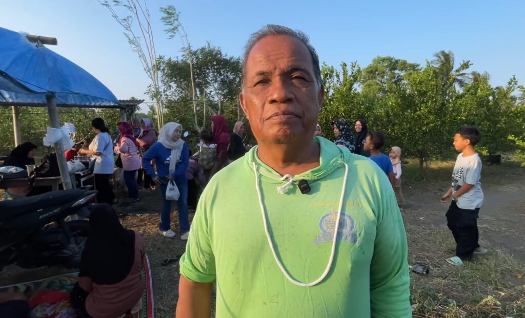 Screenshot_20260611_203441_Gallery - Mabur.co Man in a light green hoodie stands in foreground at an outdoor gathering, with a crowd and a blue tent in the background under a clear sky.