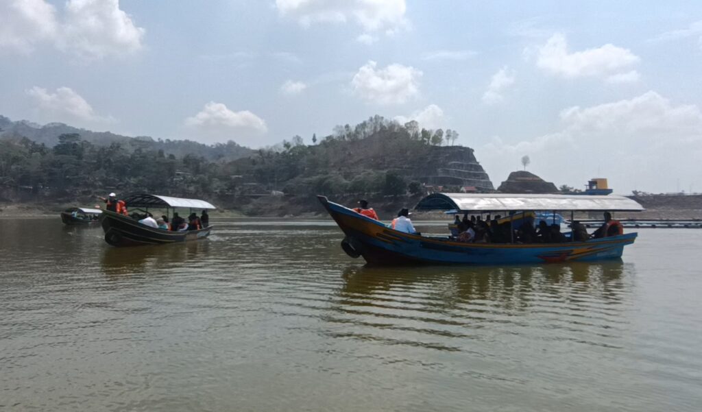 Screenshot_20260612_161307_Gallery - Mabur.co Several colorful boats with passengers in life jackets on a calm river, with hill and rocky shoreline in the background under a blue sky.