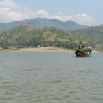 Colorful boat with passengers on a calm lake, forested hills beyond and a blue sky ahead.
