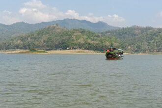Colorful boat with passengers on a calm lake, forested hills beyond and a blue sky ahead.