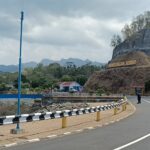 Winding road with black-and-white guard rails beside a rocky hill and a sign reading Bendungan Seromo, people walking and motorcycles nearby under a cloudy sky.