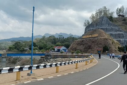 Winding road with black-and-white guard rails beside a rocky hill and a sign reading Bendungan Seromo, people walking and motorcycles nearby under a cloudy sky.