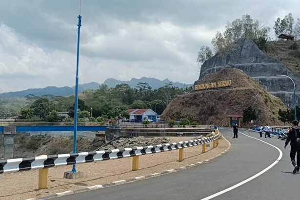 Winding road with black-and-white guard rails beside a rocky hill and a sign reading Bendungan Seromo, people walking and motorcycles nearby under a cloudy sky.