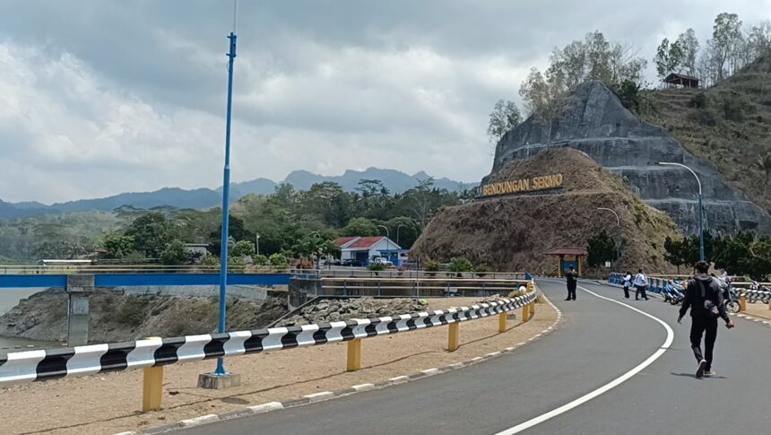 Winding road with black-and-white guard rails beside a rocky hill and a sign reading Bendungan Seromo, people walking and motorcycles nearby under a cloudy sky.