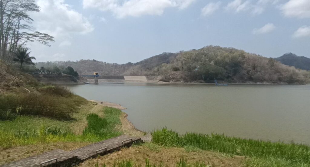 Calm lake along grassy shoreline with a small dam and a blue-and-yellow control tower in the distance, hills beyond.