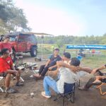 Group of men sitting in folding chairs around a small table with snacks, outdoors near parked off-road vehicles under trees.