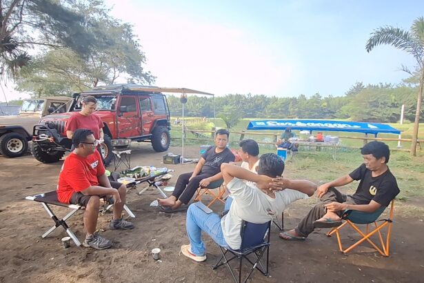 Group of men sitting in folding chairs around a small table with snacks, outdoors near parked off-road vehicles under trees.