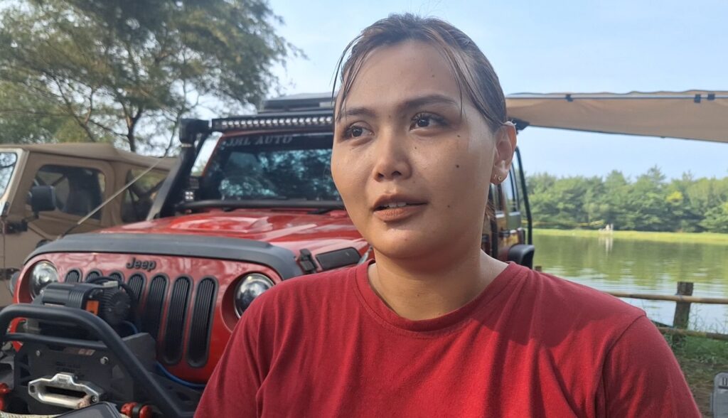 Screenshot_20260612_162409_Gallery - Mabur.co Close-up portrait of a woman in a red shirt standing beside a red Jeep by a lake on a sunny day, with trees in the background.