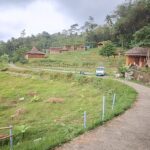 Curving concrete path through a lush hillside village with wooden huts, trees, and a parked white van nearby.