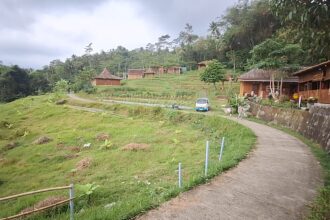 Curving concrete path through a lush hillside village with wooden huts, trees, and a parked white van nearby.