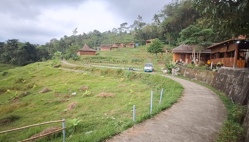 Curving concrete path through a lush hillside village with wooden huts, trees, and a parked white van nearby.