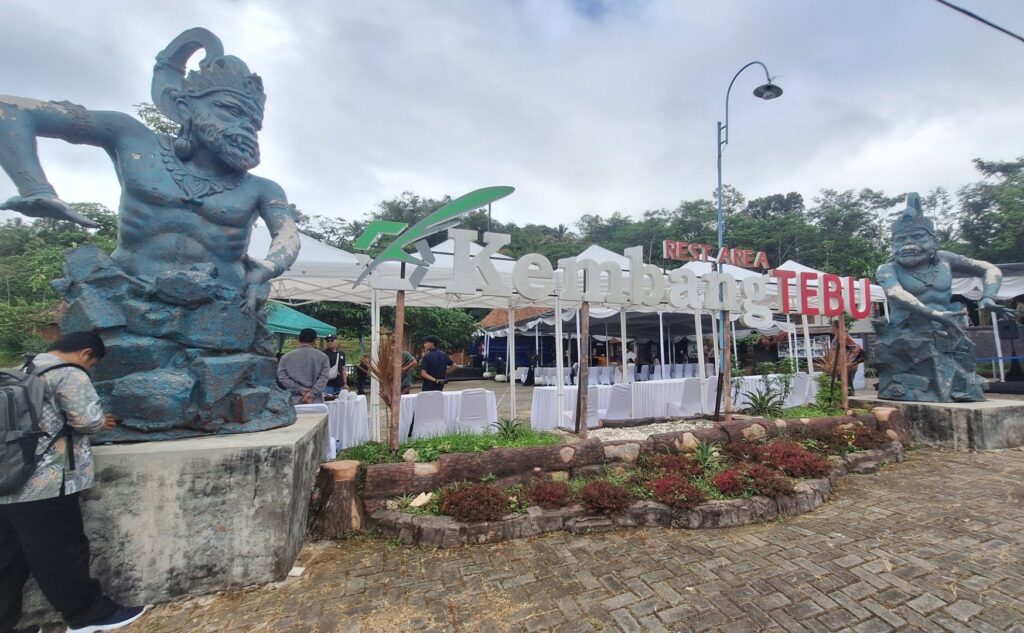 Outdoor event space with white-clothed tables under canopies, flanked by two large blue mythic statues, and a sign reading 'Kembang Tebu' Rest Area in the background.