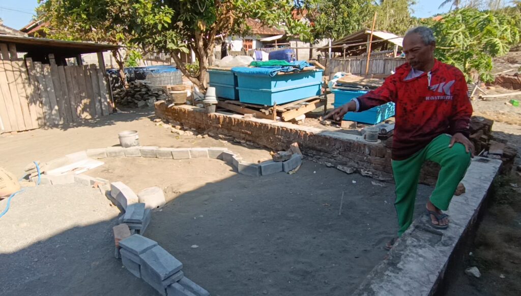 Screenshot_20260613_180715_Gallery - Mabur.co Man in a red jacket and green pants points toward a partially built circular stone wall in a yard with blue storage bins and construction materials scattered around.