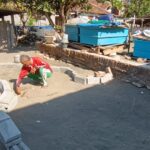 Man crouching and placing stones to form a circular garden bed or pit in a construction yard with blue crates and scattered materials nearby.