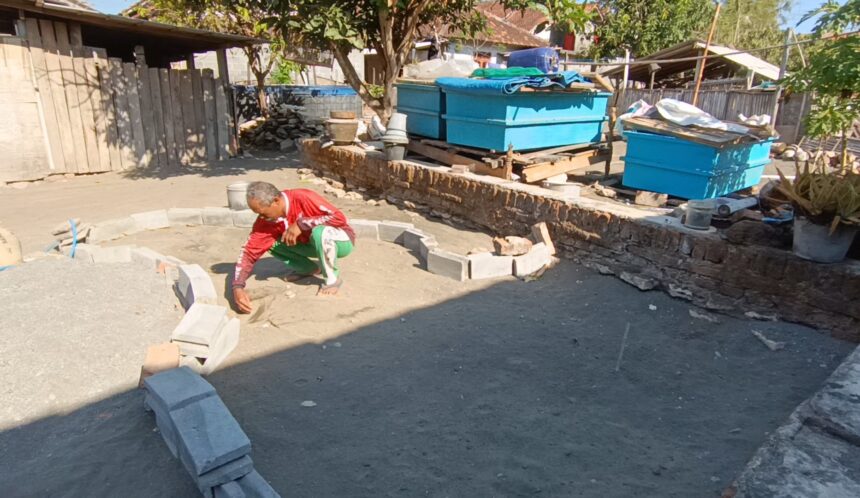 Screenshot_20260613_180741_Gallery - Mabur.co Man crouching and placing stones to form a circular garden bed or pit in a construction yard with blue crates and scattered materials nearby.