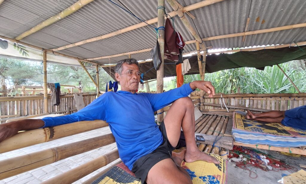 Screenshot_20260614_195813_Gallery - Mabur.co Older man in a blue long-sleeve shirt sits relaxed on a bamboo bench inside a simple shelter made of bamboo and corrugated metal.