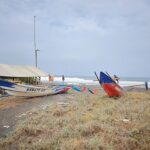 Two colorful traditional fishing boats pulled onto a dark-sand beach beside a simple shelter, with the sea and waves in the background and a cloudy sky.