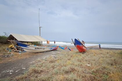 Two colorful traditional fishing boats pulled onto a dark-sand beach beside a simple shelter, with the sea and waves in the background and a cloudy sky.