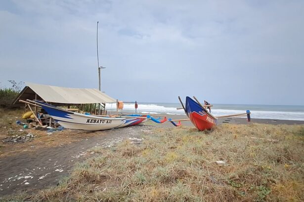 Two colorful traditional fishing boats pulled onto a dark-sand beach beside a simple shelter, with the sea and waves in the background and a cloudy sky.