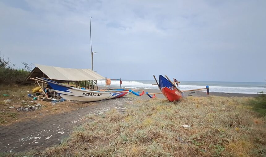 Screenshot_20260614_195848_Gallery - Mabur.co Two colorful traditional fishing boats pulled onto a dark-sand beach beside a simple shelter, with the sea and waves in the background and a cloudy sky.