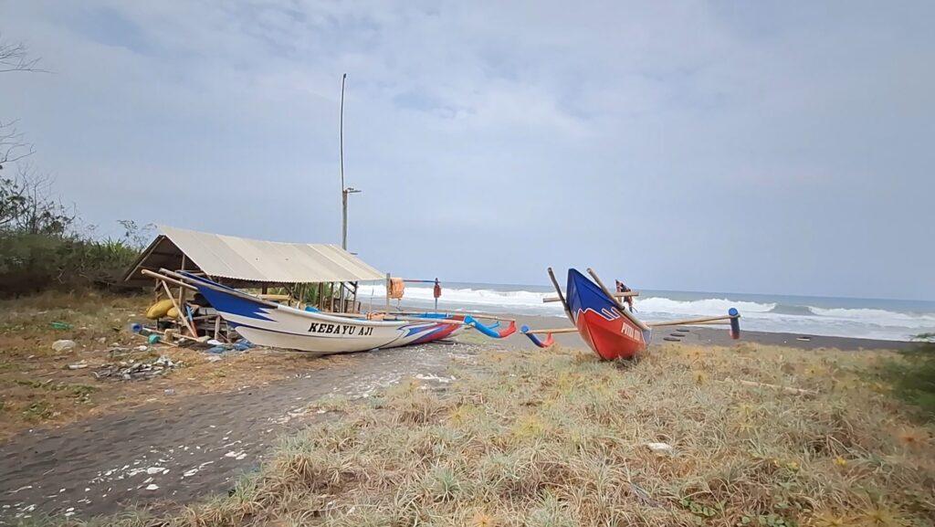 A white-and-blue fishing boat named Kebayu Aji rests on a sandy beach beside a simple shelter, with the sea in the background.