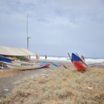 A white-and-blue fishing boat named Kebayu Aji rests on a sandy beach beside a simple shelter, with the sea in the background.