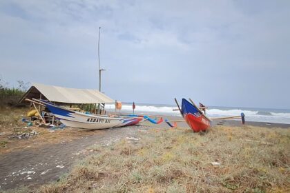 A white-and-blue fishing boat named Kebayu Aji rests on a sandy beach beside a simple shelter, with the sea in the background.