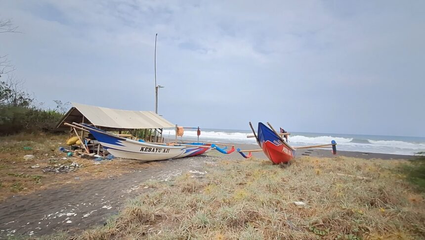 A white-and-blue fishing boat named Kebayu Aji rests on a sandy beach beside a simple shelter, with the sea in the background.