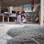 A man crouching and weaving a fishing net inside a wooden workshop, with coils of metal fencing and tools scattered around on the ground.
