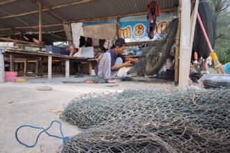 A man crouching and weaving a fishing net inside a wooden workshop, with coils of metal fencing and tools scattered around on the ground.