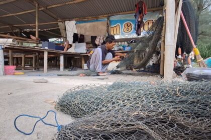 A man crouching and weaving a fishing net inside a wooden workshop, with coils of metal fencing and tools scattered around on the ground.