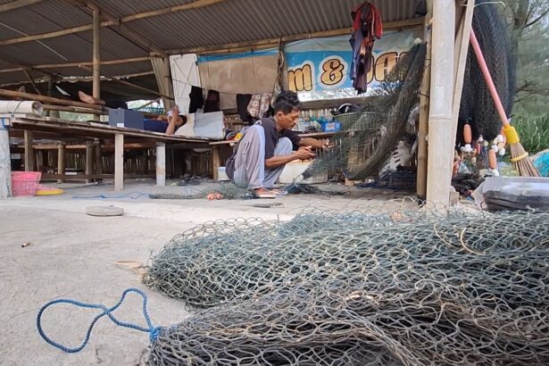A man crouching and weaving a fishing net inside a wooden workshop, with coils of metal fencing and tools scattered around on the ground.
