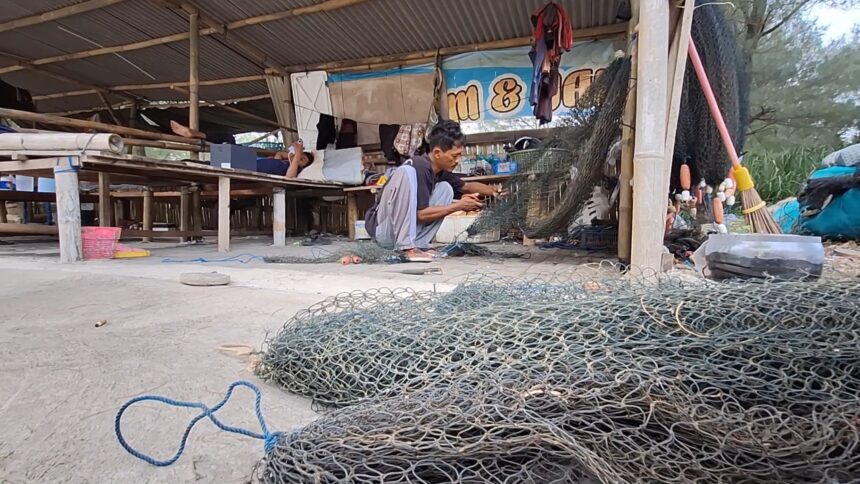 A man crouching and weaving a fishing net inside a wooden workshop, with coils of metal fencing and tools scattered around on the ground.