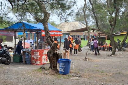Outdoor market scene with blue canopy stalls, people browsing and vendors under tents among trees in a village square