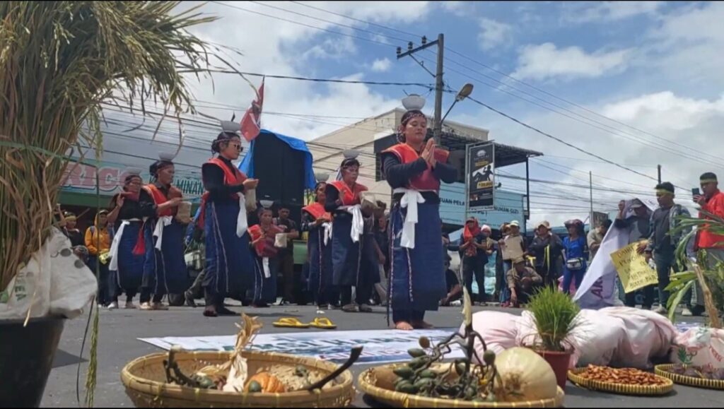 Screenshot_20260615_172911_X - Mabur.co Group of people in traditional blue and red outfits performing a street ceremony, with woven baskets of produce on the ground in the foreground and spectators watching in the background.