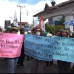 Group of protesters marching on a street, holding colorful banners and flags, with an overhead structure in the background.