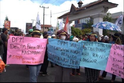 Group of protesters marching on a street, holding colorful banners and flags, with an overhead structure in the background.