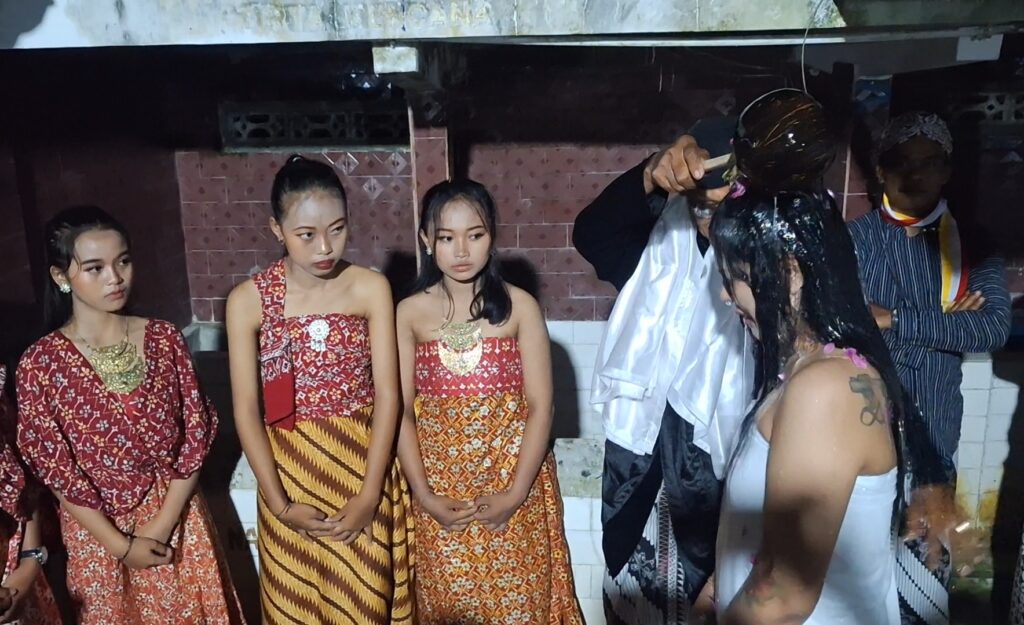 Screenshot_20260616_092214_Gallery - Mabur.co Group of four young women in matching red and gold traditional dresses stand together, as a hairstylist applies dye to another woman in white behind them backstage.
