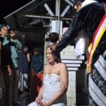 A woman in a white dress sits with eyes closed as a ceremonial hair-cutting ritual is performed by a man in a black ceremonial robe, while onlookers photograph.