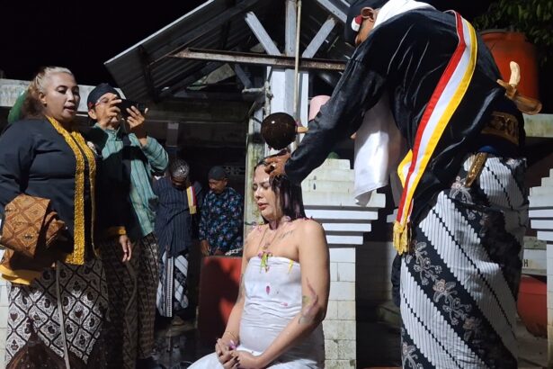 A woman in a white dress sits with eyes closed as a ceremonial hair-cutting ritual is performed by a man in a black ceremonial robe, while onlookers photograph.