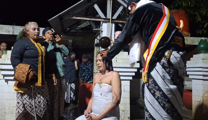 Screenshot_20260616_092306_Gallery - Mabur.co A woman in a white dress sits with eyes closed as a ceremonial hair-cutting ritual is performed by a man in a black ceremonial robe, while onlookers photograph.