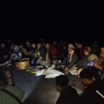Nighttime beach gathering: a group sits in a circle around ceremonial offerings and flowers on the sand. More people watch as the ceremony unfolds in dim light.