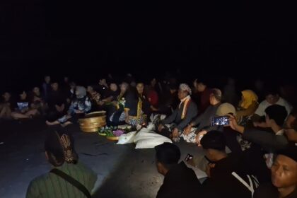 Nighttime beach gathering: a group sits in a circle around ceremonial offerings and flowers on the sand. More people watch as the ceremony unfolds in dim light.