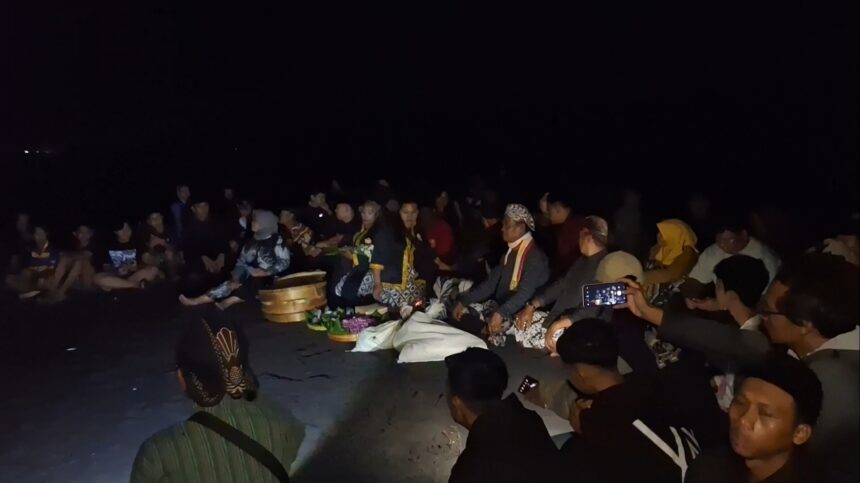 Nighttime beach gathering: a group sits in a circle around ceremonial offerings and flowers on the sand. More people watch as the ceremony unfolds in dim light.