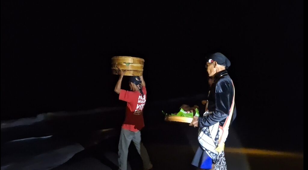 Two men walk along a dark beach at night; one wears red and lifts a large woven basket overhead, the other in blue carries a basket with green items.