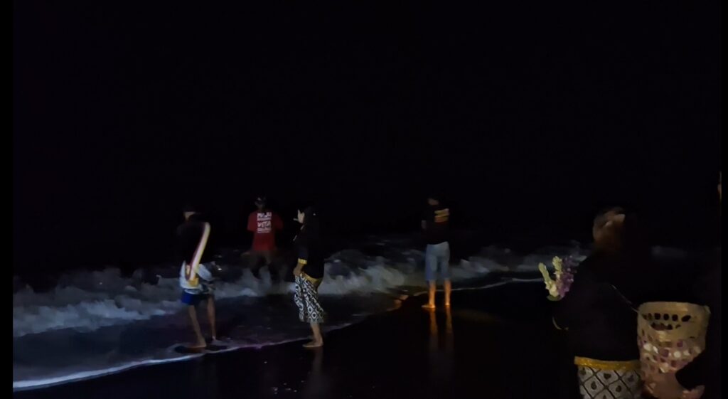 Group of people in casual clothes standing on a dark, moonlit beach as waves crash onto shore, with colorful decorative totems on the right.