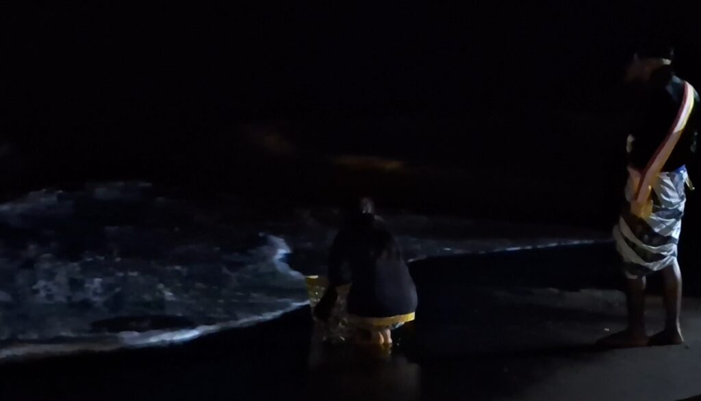 Two people stand at the edge of a dark beach at night, one crouching near the waves and the other wearing a colorful striped fabric over the shoulder.
