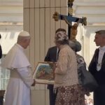 A pope-like figure in white robes receives a framed painting from a woman in a lace dress at a ceremonial gathering, with a crucifix in the background.